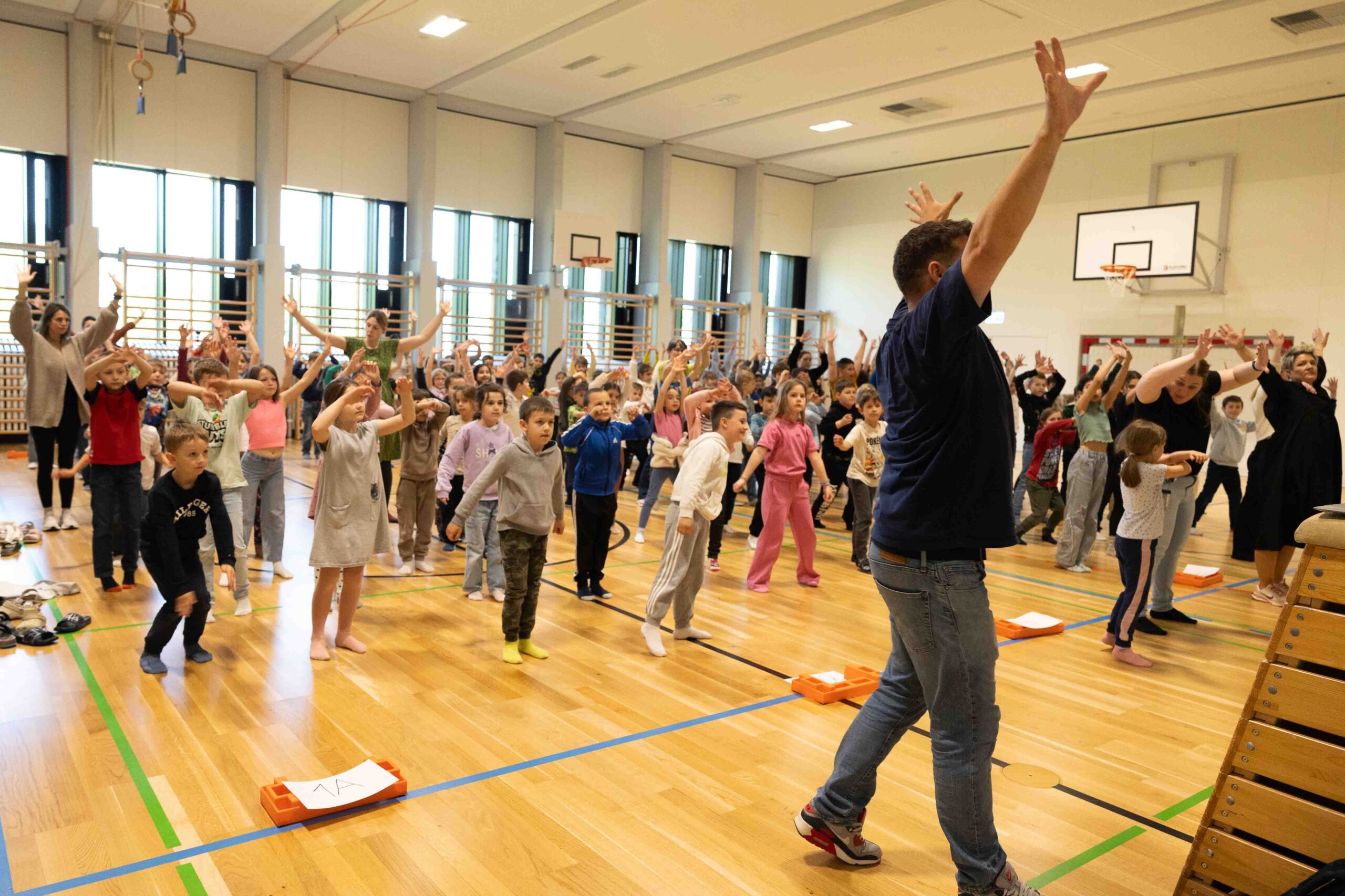 Dancing Happiness Trainer leitet eine große Gruppe Kinder und Erwachsene bei einem Live-Tanz-Workshop in einer modernen Schulturnhalle an.