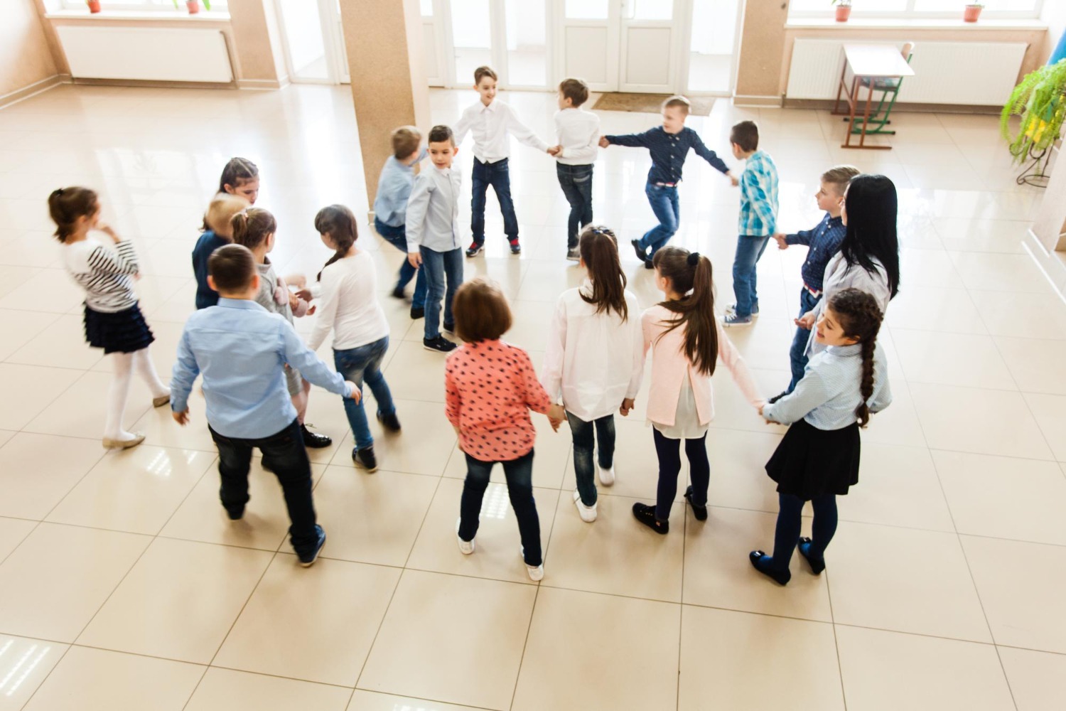 Lehrkraft zeigt Kindern Spiegel-Tanz Grundschulkinder tanzen Hand in Hand im Kreis in einem hellen Schulflur.